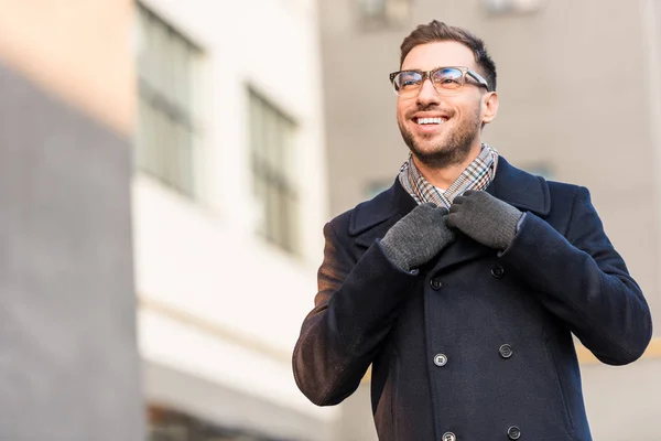 Guapo sonriente hombre sosteniendo abrigo collar con borrosa edificio - foto de stock
