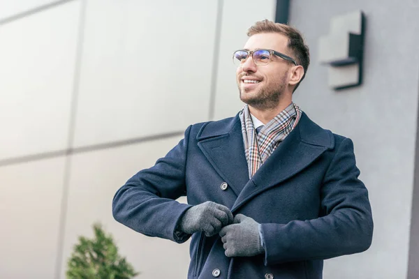 Adulte souriant bel homme en manteau de fixation de lunettes près du bâtiment — Photo de stock