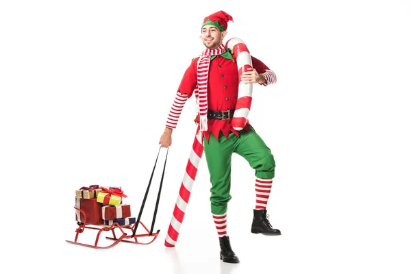 Hombre feliz en traje de elfo de Navidad llevando trineo con regalos y gran bastón de caramelo aislado en blanco - foto de stock