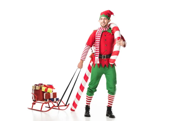 Homme heureux en costume d'elfe de Noël portant traîneau avec des cadeaux et grosse canne à bonbons isolé sur blanc — Photo de stock
