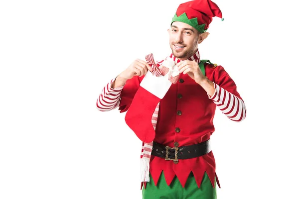 Улыбающийся мужчина в рождественском костюме эльфа кладет подарок в красный рождественский чулок и смотрит на камеру, изолированную на белом — стоковое фото
