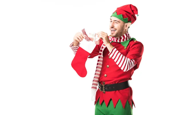 Man in christmas elf costume winging and putting present in red christmas stocking isolated on white — Stock Photo