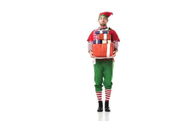 Man in christmas elf costume looking at camera and carrying pile of presents isolated on white — Stock Photo