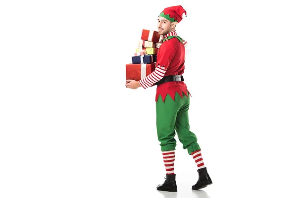 Homem feliz em traje de elfo de natal olhando para a câmera e carregando pilha de presentes isolados no branco — Fotografia de Stock