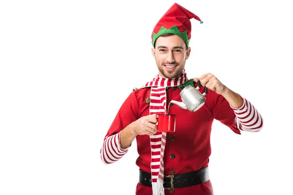 Glücklicher Mann im Weihnachtselfenkostüm mit Metallkessel, der Tee in Tasse gießt und isoliert auf weiß in die Kamera schaut — Stockfoto