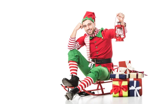 Man in christmas elf costume sitting on sleigh near pile of presents and holding red lantern isolated on white — Stock Photo