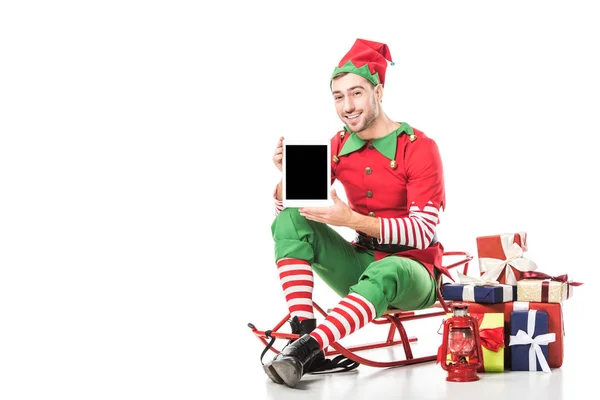 Man in christmas elf costume sitting on sleigh and holding tablet with blank screen isolated on white — Stock Photo