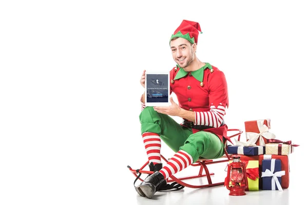 Mann im Weihnachtselfenkostüm sitzt auf Schlitten und hält Tablet mit Tumblr-App auf dem Bildschirm isoliert auf weiß — Stockfoto