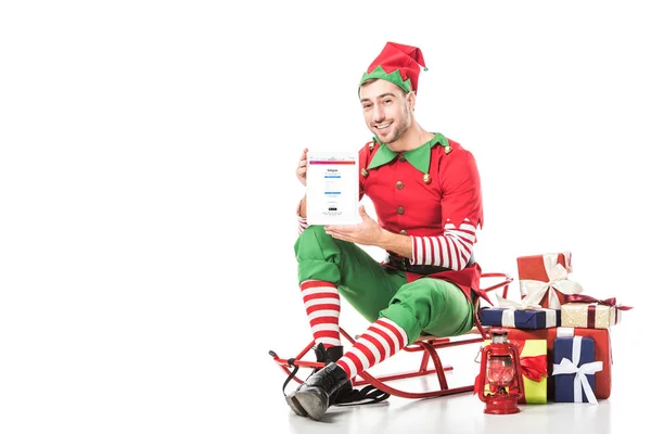 Man in christmas elf costume sitting on sleigh and holding tablet with instagram app on screen isolated on white — Stock Photo