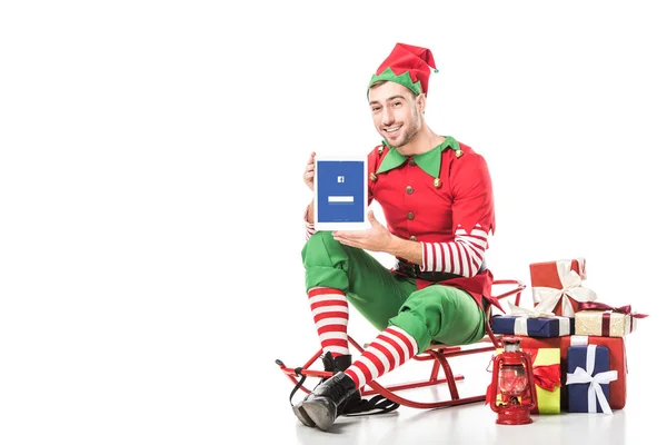 Man in christmas elf costume sitting on sleigh and holding tablet with facebook app on screen isolated on white — Stock Photo