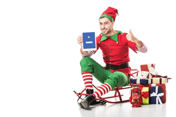 Hombre en traje de elfo de Navidad sentado en el trineo y la celebración de la tableta con aplicación de Facebook en la pantalla aislada en blanco - foto de stock