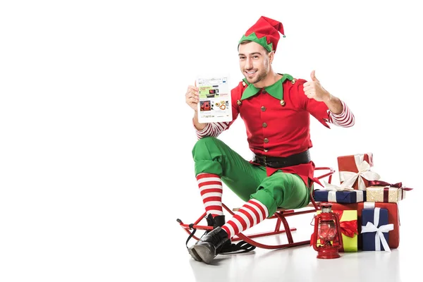 Mann im Weihnachtselfenkostüm sitzt auf Schlitten, zeigt Daumen hoch und hält Tablet mit ebay-App auf weiß — Stockfoto