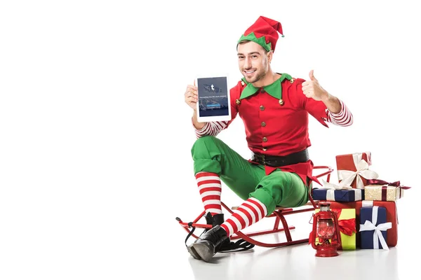 Mann im Weihnachtselfenkostüm sitzt auf Schlitten und hält Tablet mit Tumblr-App auf dem Bildschirm isoliert auf weiß — Stockfoto