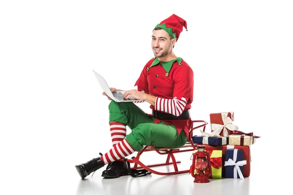 Lächelnder Mann im Weihnachtselfenkostüm sitzt auf Schlitten in der Nähe von Geschenkestapeln und benutzt Laptop isoliert auf weißem Grund — Stockfoto