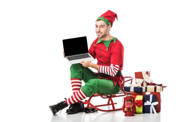 Homem em traje de elfo de natal sentado no trenó e segurando laptop com tela em branco isolado no branco — Fotografia de Stock