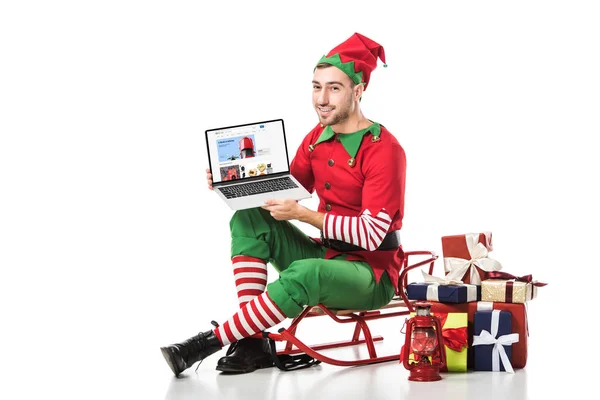 Mann im Weihnachtselfenkostüm sitzt auf Schlitten und hält Laptop mit ebay-Website auf dem Bildschirm isoliert auf weiß — Stockfoto
