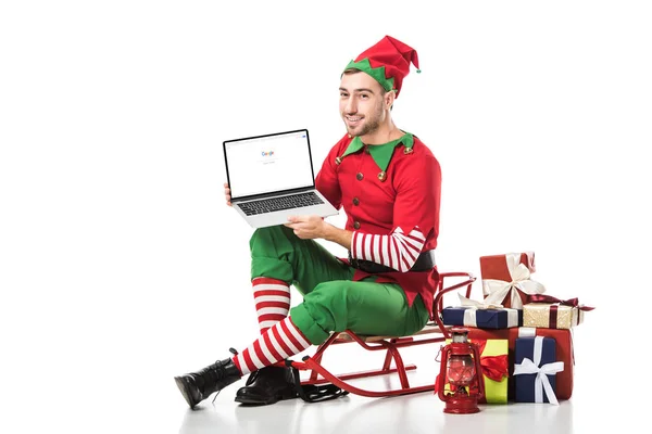 Hombre en traje de elfo de Navidad sentado en el trineo y la celebración de la computadora portátil con el sitio web de Google en la pantalla aislada en blanco - foto de stock