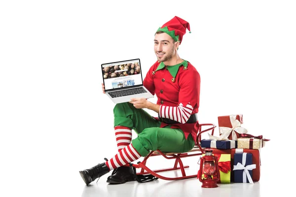 Man in christmas elf costume sitting on sleigh and holding laptop with depositphotos website on screen isolated on white — Stock Photo