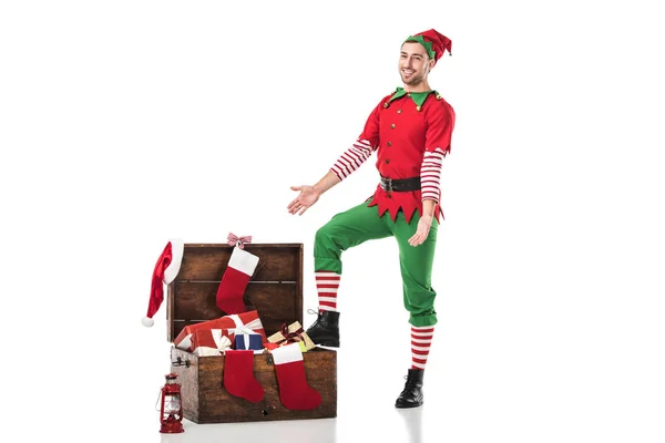 Lächelnder Mann im Weihnachtselfenkostüm, der neben einer Holzkiste voller Geschenke steht, mit ausgestreckten Händen auf weißem Grund. — Stockfoto