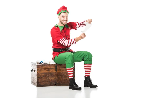 Весёлый мужчина в рождественском костюме эльфа сидит на деревянном сундуке и держит список желаний изолированным на белом — стоковое фото