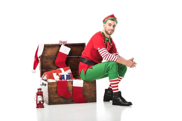 Hombre en traje de elfo de Navidad sentado en el pecho de madera con medias de Navidad y regalos aislados en blanco - foto de stock