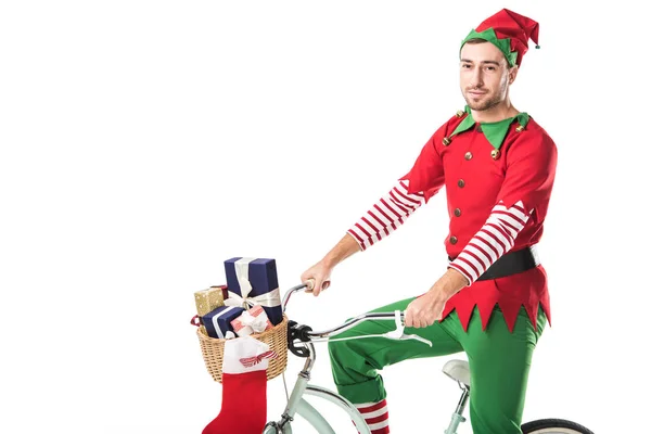 Uomo in costume elfo di Natale guardando la macchina fotografica e in sella alla bicicletta con cestino pieno di regali isolati su bianco — Foto stock