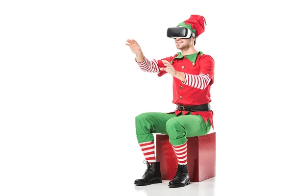 Man in christmas elf costume sitting on box and wearing virtual reality headset isolated on white — Stock Photo