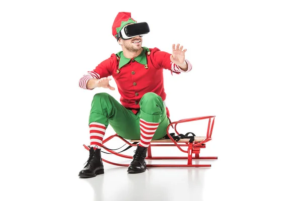 Mann im Weihnachtselfenkostüm sitzt auf Schlitten und trägt Virtual-Reality-Headset isoliert auf weißem Grund — Stockfoto