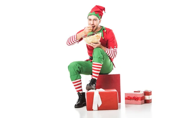 Hombre feliz en traje de elfo de Navidad sentado en un montón de regalos y comer palomitas de maíz aislado en blanco - foto de stock