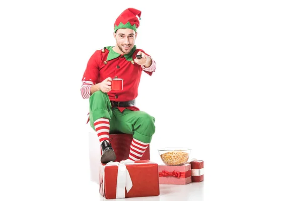 Hombre en traje de elfo de Navidad sentado en un montón de regalos, sosteniendo la taza de té y apuntando control remoto a la cámara aislada en blanco - foto de stock