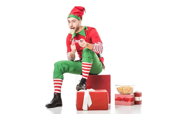 Hombre en traje de elfo de Navidad sentado en un montón de regalos con piruleta y jugando videojuego aislado en blanco - foto de stock