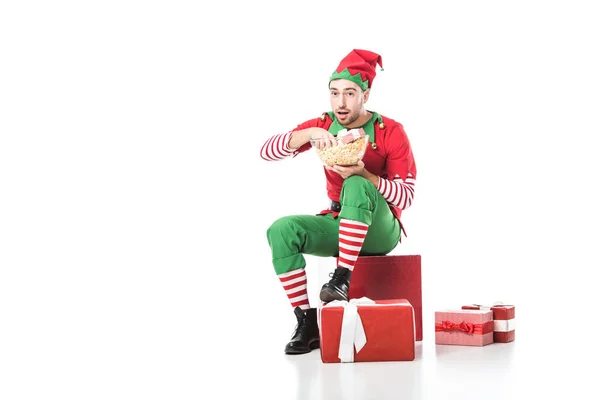 Hombre en traje de elfo de Navidad sentado en un montón de regalos y comer palomitas de maíz aislado en blanco - foto de stock