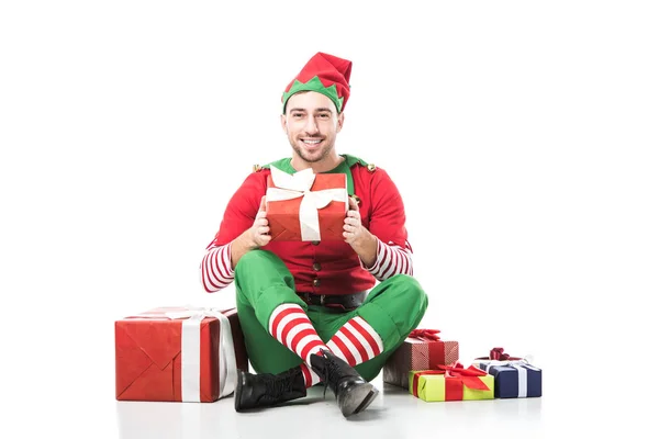Glücklich lächelnder Mann im Weihnachtselfenkostüm, der neben einem Stapel Geschenke sitzt und Geschenke isoliert auf weiß hält — Stockfoto