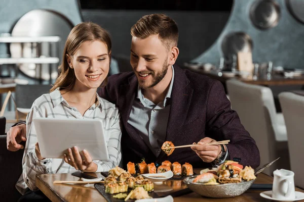 Attractive couple using digital tablet while eating sushi in restaurant — Stock Photo