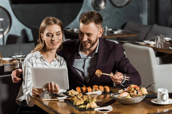 Attractive couple of yound adult using digital tablet while eating sushi in restaurant — Stock Photo