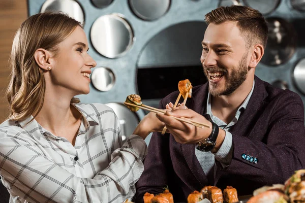 Glückliches lächelndes Paar junger Erwachsener, die sich im Restaurant gegenseitig mit Sushi füttern — Stockfoto