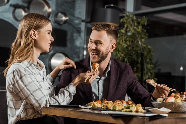 Красива усміхнена молода жінка годує свого хлопця суші в ресторані — стокове фото