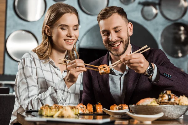 Souriant attrayant jeune couple adulte mangeant des sushis ensemble au restaurant — Photo de stock