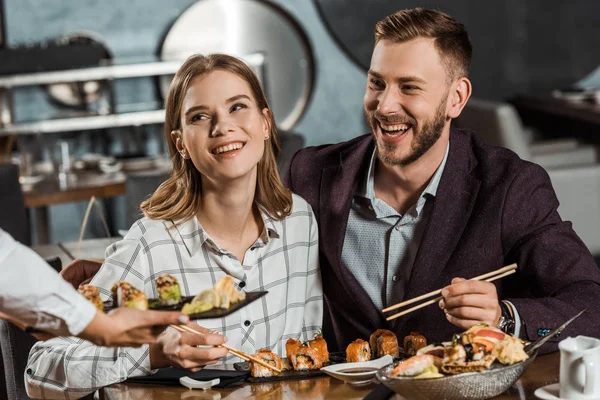 Pareja sonriente comiendo rollos de sushi mientras el camarero trae un nuevo orden en el restaurante - foto de stock