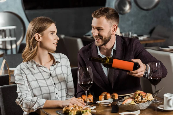 Happy young adult couple having dinner while man pouring wine in glasses in restaurant — Stock Photo