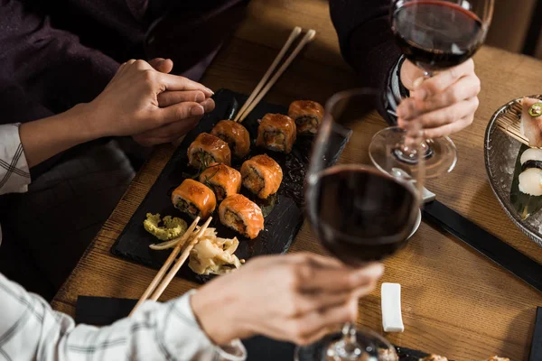 Vue partielle du couple se tenant la main tout en buvant du vin et en mangeant des sushis au restaurant — Photo de stock