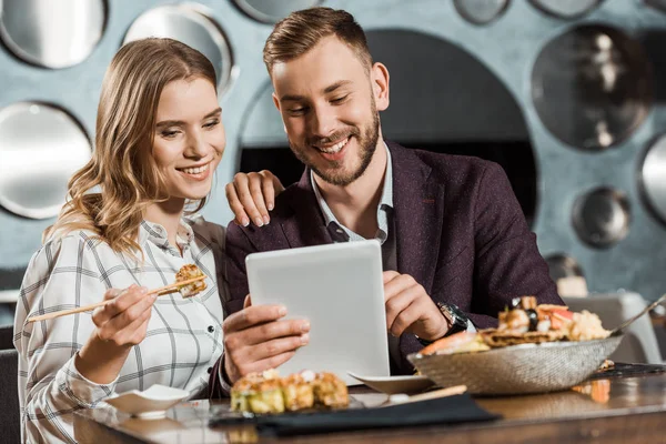 Atractiva y sonriente pareja de jóvenes adultos usando tableta digital mientras cenan en el restaurante - foto de stock