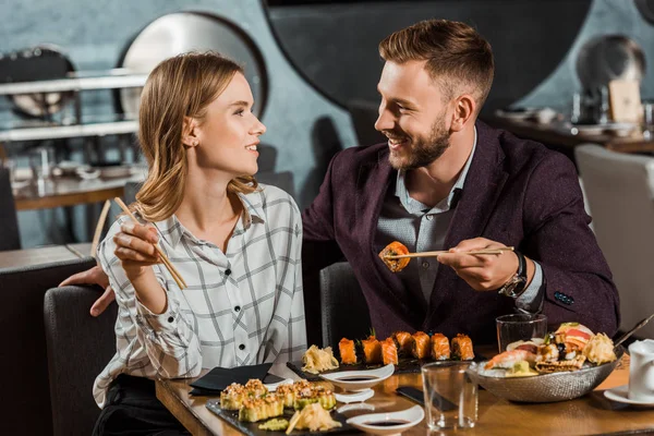 Feliz jovem casal adulto olhando um para o outro enquanto comendo rolos de sushi no restaurante — Fotografia de Stock
