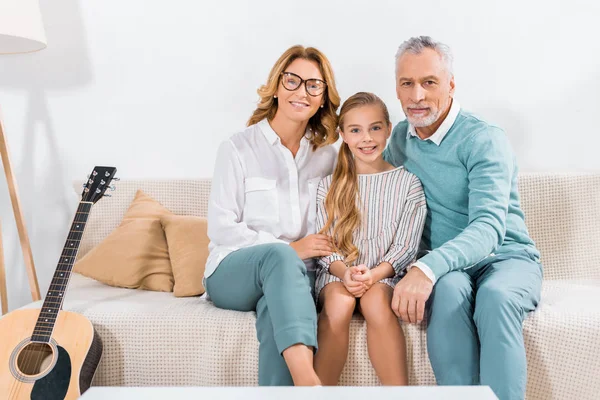 Grands-parents embrassant petite-fille souriante et regardant la caméra sur le canapé à la maison — Photo de stock