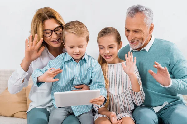 Abuelos y nietos saludando con las manos mientras hacen videollamadas con tabletas digitales en casa - foto de stock