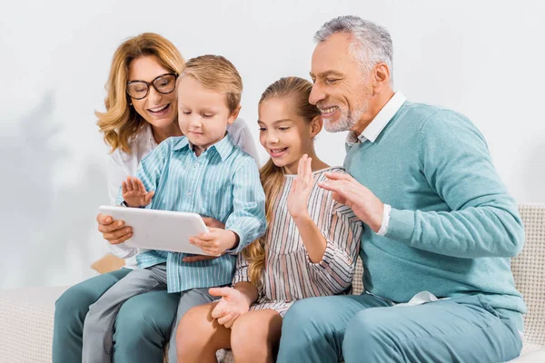 Familia feliz saludando con las manos mientras tiene videollamada con tableta digital en casa - foto de stock