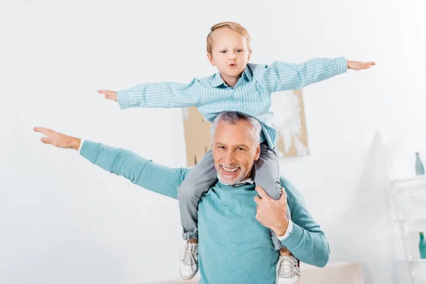 Child sitting on happy grandfather shoulders and imitating plane at home — Stock Photo