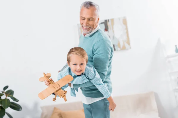 Mature man holding on hands grandson with wooden airplane and having fun at home — Stock Photo