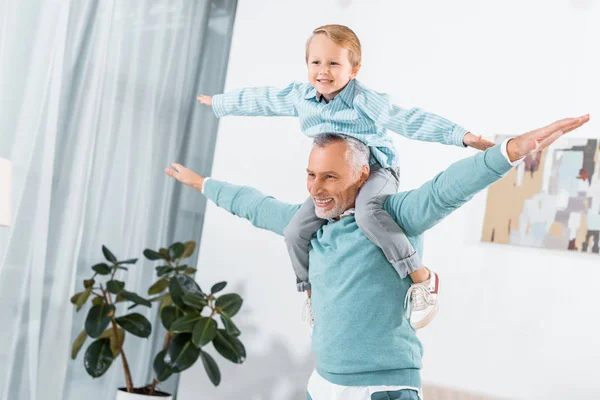 Смеющийся малыш сидит на плечах дедушки и веселится дома. — стоковое фото