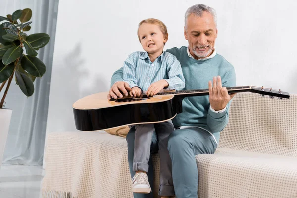 Allegro nonno che gioca con il nipote in ginocchio suonando la chitarra acustica a casa — Foto stock